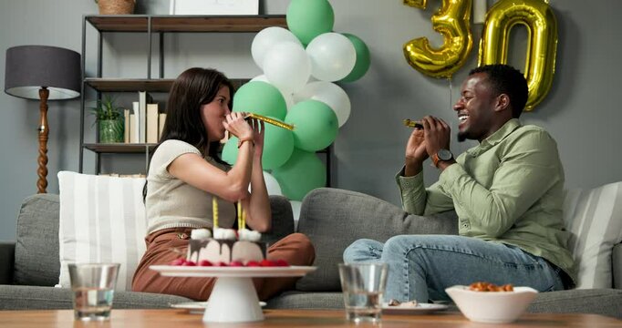 Happy multiracial and african american couple sitting on sofa, blowing in birthday trumpets at a bir