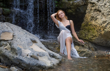 A girl in a transparent white dress sits on the rocks by the waterfall
