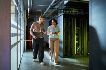 Male and female colleagues engaged in conversation while walking through an IT server room with...
