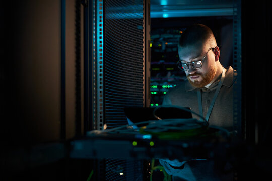 Technician in server room inspecting hardware components of data center equipment, surrounded by servers and cables, engrossed in work while holding tablet for diagnostics