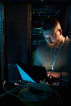 Technician working in dimly lit server room, analyzing data on laptop. Surrounded by networking cables and switches, focusing on tasks without direct eye contact with camera