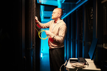 Fototapeta na wymiar Technician connecting yellow network cables in modern server room while standing near computer and other equipment, with blue lighting in background
