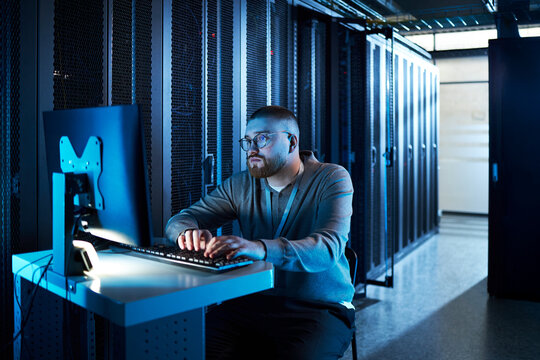 Man sitting in data center and working on computer, surrounded by server racks. He is focused and engaged in his task, creating a tech-centric atmosphere