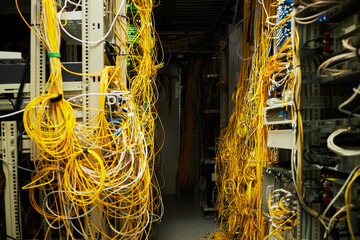 A dense cluster of yellow network cables entangling through various server racks in a busy server room illuminated by artificial lighting. Visible terminals and routing hardware add to the complexity