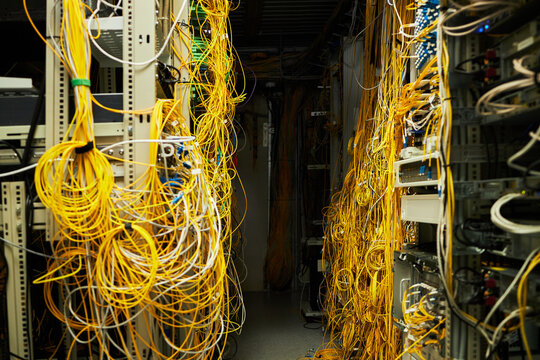 A dense cluster of yellow network cables entangling through various server racks in a busy server room illuminated by artificial lighting. Visible terminals and routing hardware add to the complexity