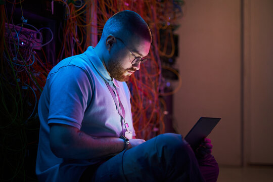 Technician working in dimly lit server room with colorful network cables using a digital tablet for troubleshooting and maintenance tasks