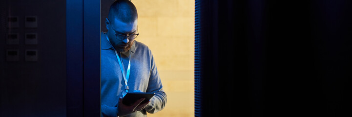 Man standing in dimly lit room with blue light, holding and engaging with tablet device while...