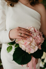 A bride in a white dress on her wedding day holds a bouquet of pink roses and strokes it with her hand with a gold wedding ring on her finger