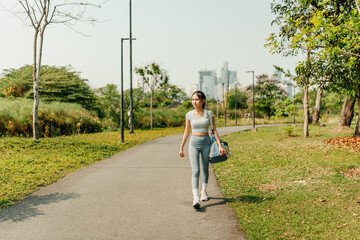 A woman is walking along a paved path in a green urban park, dressed in athletic wear including...
