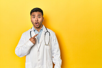 Asian doctor in white coat with stethoscope, yellow studio pointing to the side