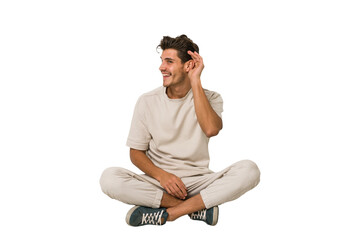 Young caucasian man sitting on the floor isolated on white background trying to listening a gossip.