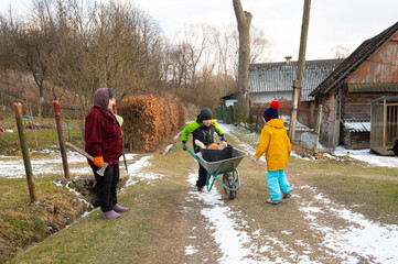 Children playing in the countryside during winter while helping each other with a wheelbarrow on a snowy path surrounded by nature and rustic buildings