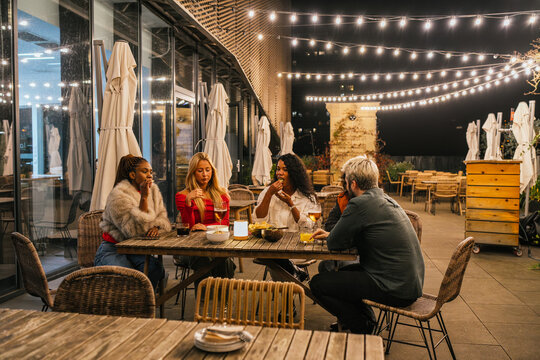 Multiethnic group of friends enjoying drinks and food at a rooftop party, illuminated by warm string lights, creating a cozy and festive atmosphere