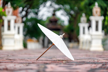 white paper umbrella, a traditional Lanna craft, stands on a textured surface in San Sai, Chiang Mai, Thailand, with blurred temple structures in the background.