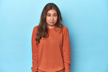 Teen girl in vibrant orange sweater on blue shrugs shoulders and open eyes confused.