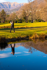 Male Golfer Reflected in a Water Pond and Hitting the Golf Ball on Fairway on Golf Course with Mountain in a Sunny Day in Switzerland.