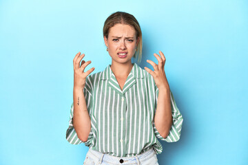 Blonde young woman in green striped shirt on blue background upset screaming with tense hands.