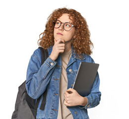 Young Caucasian redhead student with books looking sideways with doubtful and skeptical expression.