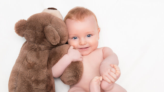 A modestly smiling baby lies hugging a teddy bear and looks at the camera
