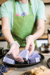 Woman in green apron presenting freshly crafted lavender soap bars on a plate with gloved hands. Handmade wellness, chemical-free skincare, female entrepreneurship, botanical beauty
