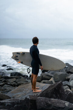 Man with surfboard standing on rock against cloudy sky