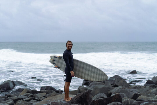Man with surfboard standing on rock against cloudy sky