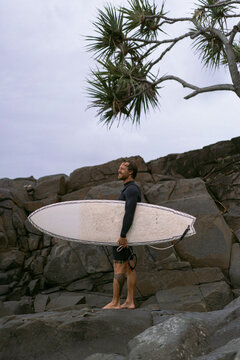 Man with surfboard standing on rock against cloudy sky