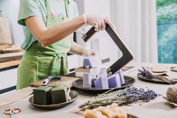 Woman in green apron slicing handmade lavender soap on a worktable with bunches of dried lavender nearby. Clean beauty, natural ingredients, chemical-free soap, small-scale production
