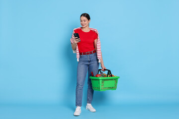 Smiling woman with basket full of products and smartphone on light blue background. Online shopping