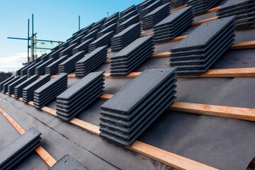 Construction worker carefully places roof tiles on a sloped surface while standing on top of a building under a clear blue sky, showcasing the effort involved in roofing work
