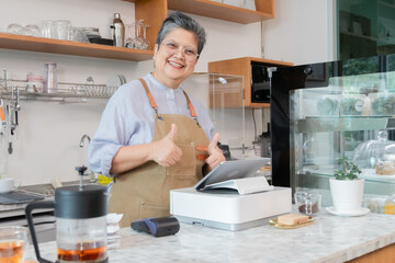 Senior entrepreneur cafe owner barista hold POS machine to customer, Asian female in apron welcome warmly in counter, croissants and glassware background, retired elderly woman start small business