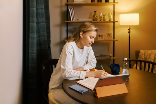 Woman writing in a notebook while sitting at a table, indoor setting during the evening
