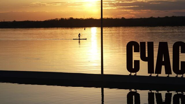 Golden sunset illuminates calm lake water as man paddles on stand up paddleboard, with large cyrillic letters spelling happiness in foreground