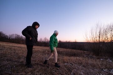 Two friends explore a serene field at dusk, wrapped in warm layers as day transitions to night,...