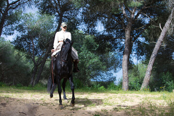 Young and beautiful woman riding a purebred Spanish black horse in a pine forest. The woman is dressed in typical horsewoman costume. In the background the lush vegetation.