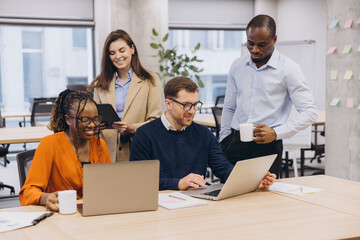 Multiracial business team working together in a contemporary office, using laptops and discussing ideas over coffee. Teamwork and collaboration emphasized