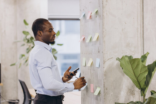 Focused african american businessman holding a smartphone and organizing his work using sticky notes on a wall in the office - Powered by Adobe