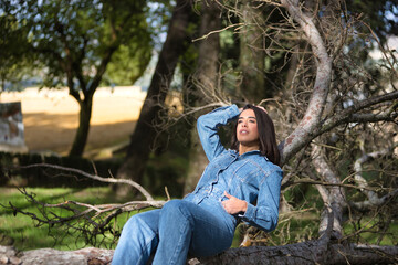 Fototapeta premium Young, beautiful, brunette, Hispanic woman, wearing a denim suit, pensive and distracted, lying on the fallen trunk of a tree, in nature, receiving the sun's rays. Concept fashion, beauty, femininity.