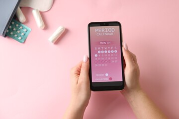 Menstruation. Woman holding smartphone with period calendar application on pink background, top view
