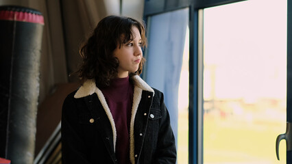 A thoughtful girl by a window looks outward, framed by a warm and sunny background. This image symbolizes personal reflection, peacefulness, and connection with the world.
