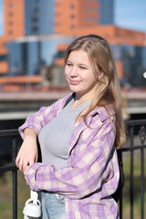 Fototapeta premium A teenage girl enjoys the sunlight as she poses by a railing, holding headphones in her hand. The photo captures the vibrancy and freedom of youth in an urban setting.
