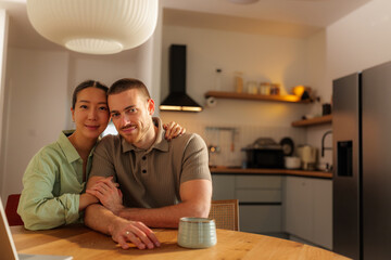 Young multi ethnic couple sitting at a kitchen table, embracing and sharing smiles, enjoying...