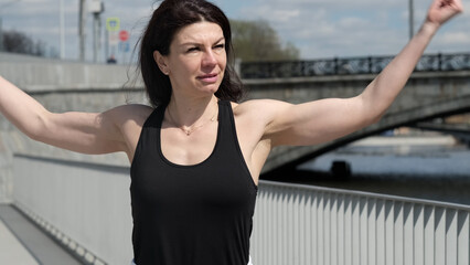 A determined woman in a black tank top stretches her arms wide in an urban setting, symbolizing...
