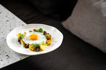 A gourmet dish with a sunny-side-up egg, fresh vegetables, pickles, mushrooms, and herbs, artfully presented on a white plate against a stylish, modern countertop. Vibrant colors and textures enhance 
