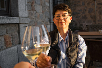 Smiling senior woman toasting with glasses of white wine on an outdoor terrace, enjoying the warm afternoon sunlight and celebrating a special moment.