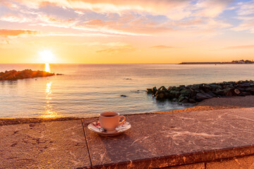 Obraz premium cup of coffee or tea on a morning embarkment beach with blue sea and beautiful cloudy sunrise or sunset on background, street food breakfast concept
