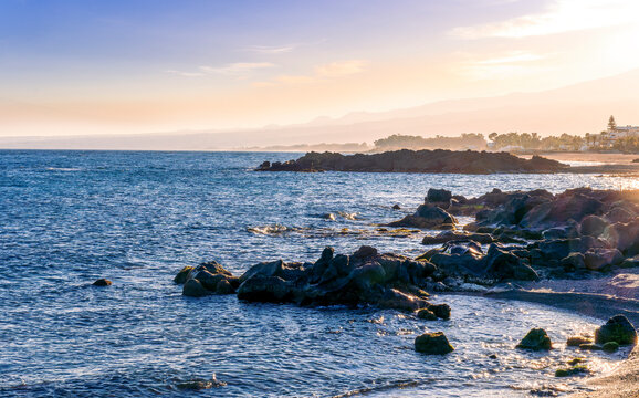 black rock sea beach during sunrise or sunset with golden sand, mild surf, blue calm water and beautiful cloudy sky with coast and mountains on background