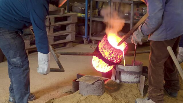 Artisans carefully handle glowing crucible as molten bronze flows into sand mold during traditional bell production using floor casting methods, slow motion