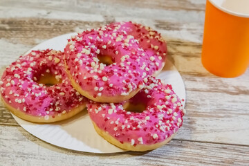 Pink doughnuts with sprinkles on a wooden kitchen table next to an orange glass.