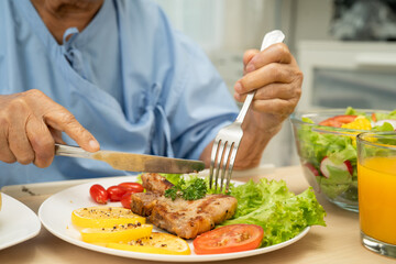 Asian senior woman patient eating pork chop stake and vegetable salad for healthy food in hospital.